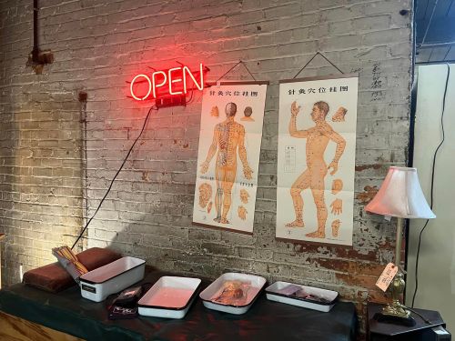 A brick wall with an "OPEN" neon sign, two anatomical charts, and a table with trays and a lamp.