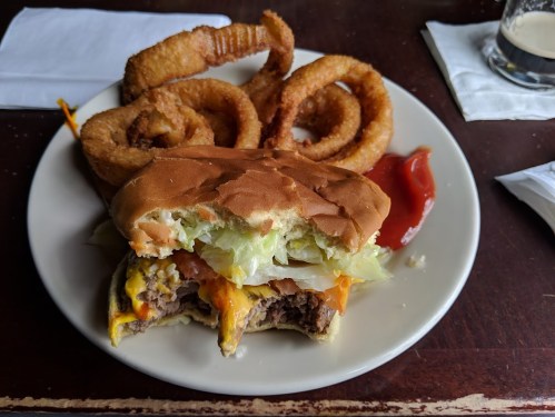 A plate with a half-eaten cheeseburger topped with lettuce and tomato, alongside crispy onion rings and ketchup.