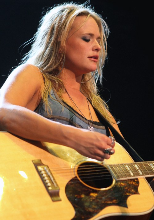 A female musician plays an acoustic guitar, eyes closed, with a focused expression and long, wavy hair.