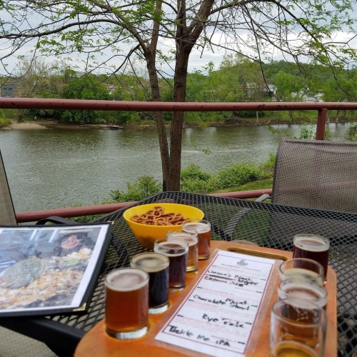 A wooden flight of craft beers sits on a table by a river, with snacks and a scenic view of trees and water.