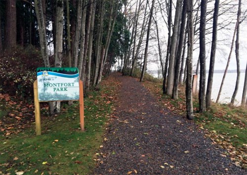 A gravel path lined with trees leads to a sign for Montfort Park near a body of water.