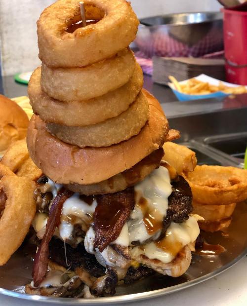 A towering burger topped with onion rings, bacon, and melted cheese, surrounded by more onion rings and fries.