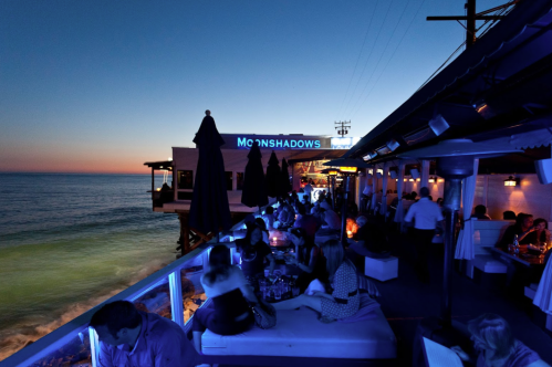A lively seaside restaurant at dusk, with people dining and enjoying the ocean view under blue lighting.