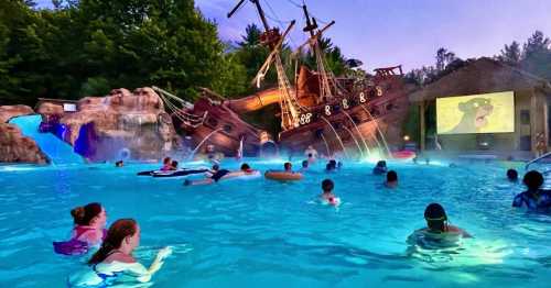 A lively pool scene at dusk with a pirate ship structure, people swimming, and a movie projected on a screen.