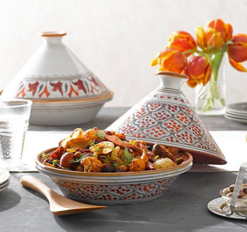 A colorful dish of roasted vegetables in a decorative bowl, with a tagine and flowers in the background.