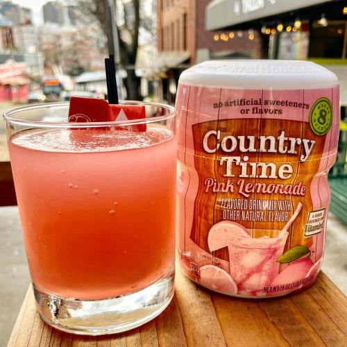 A glass of pink lemonade with a straw next to a container of Country Time Pink Lemonade mix on a wooden surface.