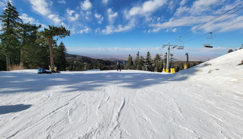 A snowy ski slope with a clear blue sky, trees in the background, and ski lifts in the distance.