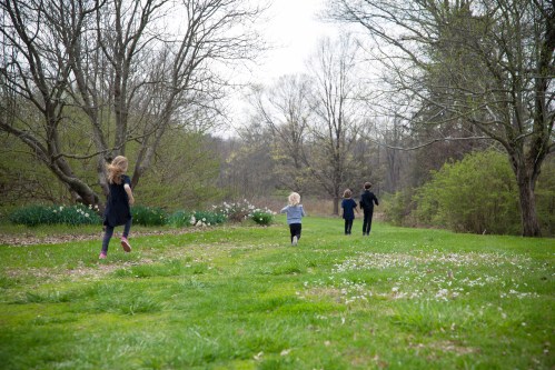 Four children running through a grassy field, surrounded by trees and blooming flowers on a cloudy day.