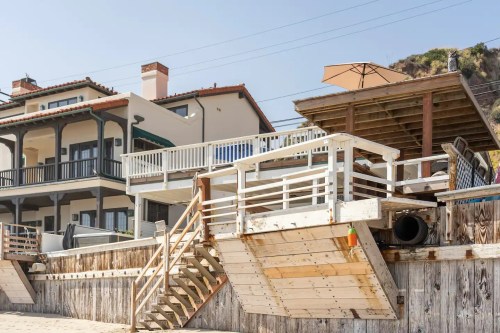 A beachside house with a deck, stairs, and an umbrella, overlooking a sandy area and wooden fencing.