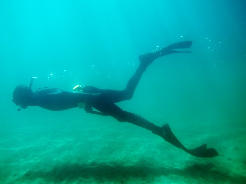 A diver swims underwater, wearing fins and a snorkel, surrounded by blue water and bubbles.