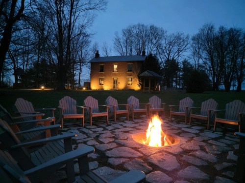 A cozy outdoor fire pit surrounded by wooden chairs, with a house illuminated in the background at dusk.