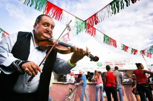 A man plays the violin at a festive outdoor event, with colorful banners and food stalls in the background.