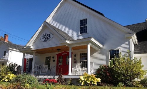 A white house with a red door and porch, surrounded by greenery and blue sky.