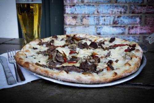 A pizza topped with beef, peppers, and cheese, served with a glass of beer on a wooden table.