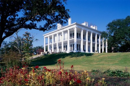 A grand white mansion with columns, surrounded by green grass and colorful flowers under a clear blue sky.