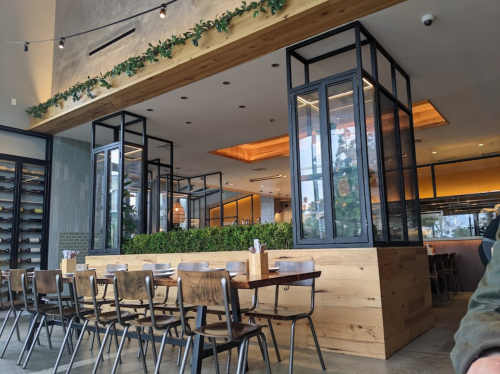 Modern restaurant interior featuring wooden tables, metal chairs, and greenery, with large windows and warm lighting.