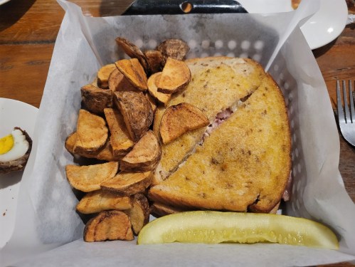 A sandwich cut in half, served with crispy potato wedges and a pickle slice, in a lined basket.