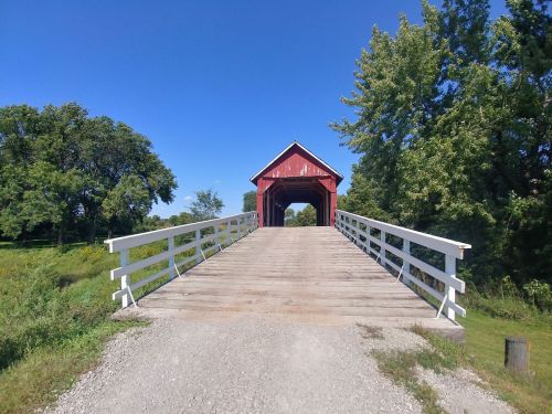 A red covered bridge with a wooden walkway, surrounded by green trees and a clear blue sky.