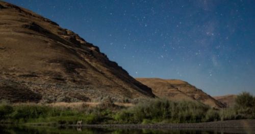 A serene landscape at night featuring a river, hills, and a starry sky.