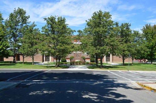 A view of a brick building surrounded by trees and a clear blue sky, with pathways and grassy areas in the foreground.