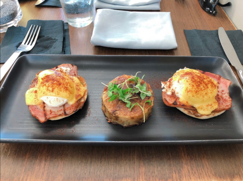 A black plate with two eggs Benedict topped with ham and hollandaise sauce, and a potato cake garnished with greens.