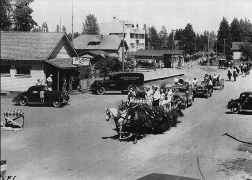 A historic parade with horse-drawn carriages, vintage cars, and people in period clothing on a dirt road.