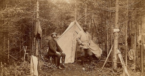 Two men in vintage clothing stand outside a tent in a forest, surrounded by trees and camping gear.