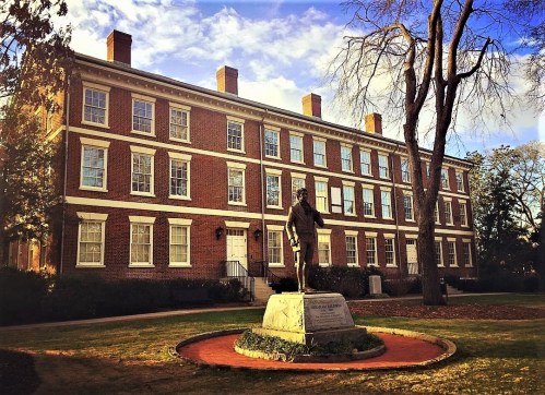 A brick building with large windows and a statue in front, set against a blue sky with scattered clouds.