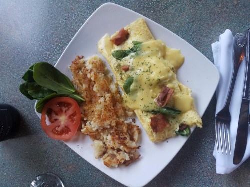A plate with an omelette topped with cheese and spinach, hash browns, a slice of tomato, and a side of spinach.