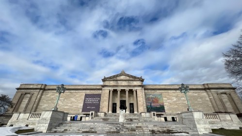 A grand museum building with a stone facade, steps, and sculptures, set against a cloudy sky. Snow is visible in the foreground.