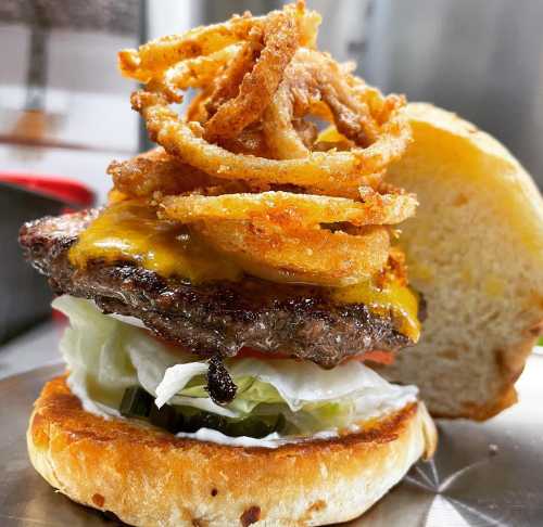 A close-up of a juicy cheeseburger topped with crispy onion rings, lettuce, tomato, and pickles on a toasted bun.