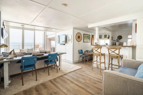 Bright coastal living space with a dining area, kitchen bar, and large windows overlooking the ocean. Cozy and inviting decor.