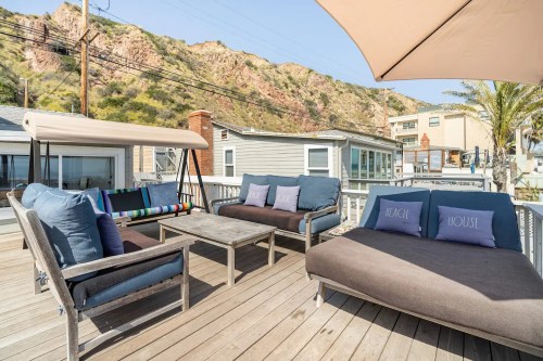 A cozy outdoor seating area with plush couches and a table, surrounded by hills and beach houses.