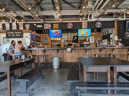 A bar with a wooden counter, stools, and tables; patrons seated, screens displaying images, and various decor on the walls.