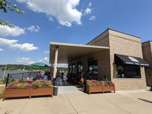 Exterior of Green Tree Brewery with outdoor seating, colorful flower planters, and a view of the water under a blue sky.