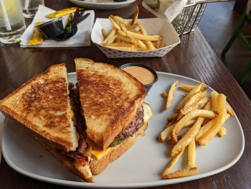 A grilled sandwich with a beef patty and cheese, served with a side of fries and dipping sauce on a white plate.