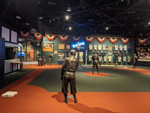 A museum exhibit featuring life-sized bronze statues of baseball players in a green field setting, with memorabilia on display.