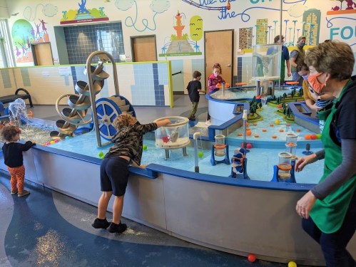 Children play at a water activity station, exploring water features and colorful balls, with adults supervising nearby.