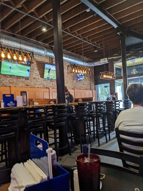 Interior of a restaurant with wooden beams, bar seating, and TVs showing sports. A few patrons are seated.