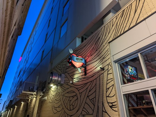 A building exterior featuring a colorful "MOM" sign and artistic wall mural, illuminated at dusk.