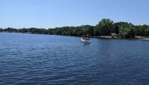 A small boat on a calm lake surrounded by green trees under a clear blue sky.