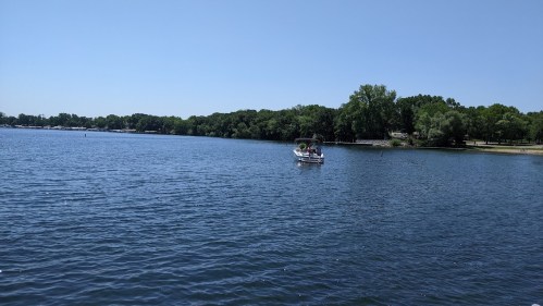 A calm lake scene with a boat on the water and lush green trees lining the shore under a clear blue sky.