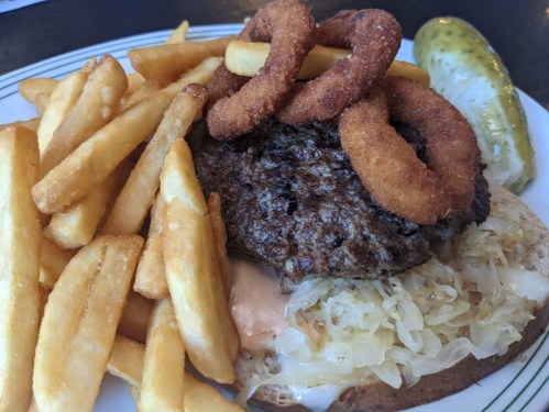 A plate featuring a burger with onion rings, sauerkraut, and fries, alongside a pickle.