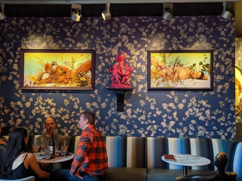 A vibrant restaurant interior with floral wallpaper, two framed artworks, and a sculpture, featuring diners at a table.