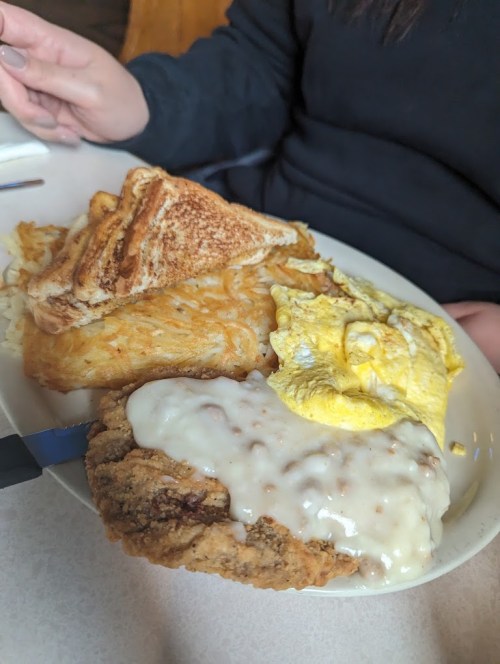 A plate of breakfast featuring fried chicken, gravy, scrambled eggs, hash browns, and toast.