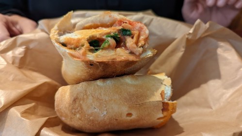 A close-up of a halved sandwich filled with tomato, cheese, and herbs, resting on crumpled brown paper.