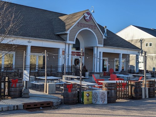 A cozy restaurant with outdoor seating, festive decorations, and barrels, set against a clear blue sky.