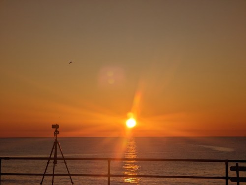 A sunset over the ocean, with a camera on a tripod in the foreground capturing the vibrant colors.
