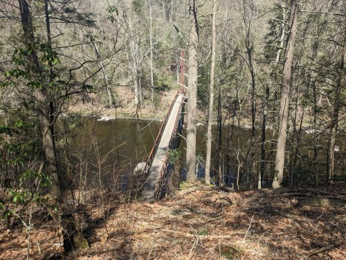 A narrow suspension bridge spans a calm river, surrounded by bare trees and rocky banks in a wooded area.