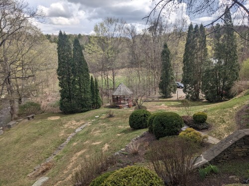 A serene landscape featuring a gazebo surrounded by trees and shrubs, with a grassy area and distant hills.